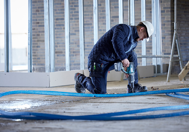 Handwerker in Arbeitskleidung und Schutzhelm kniet auf einer Baustelle und bohrt mit einer elektrischen Maschine in den Boden.