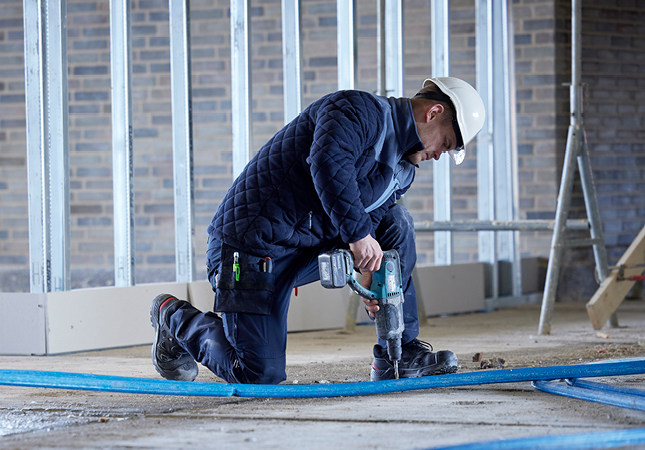 Handwerker in Arbeitskleidung und Schutzhelm kniet auf einer Baustelle und bohrt mit einer elektrischen Maschine in den Boden.