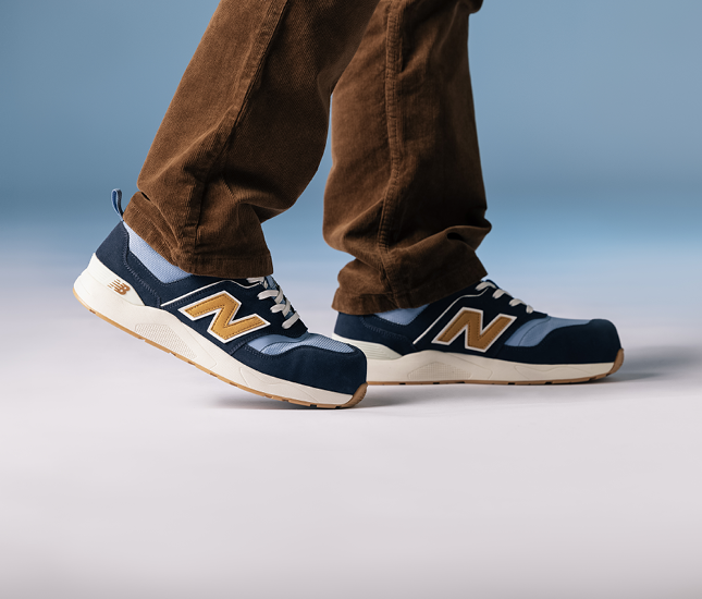 Ein Paar New Balance Sneakers in Blau und Orange, getragen von einer Person mit braunen Cordhosen. Die Schuhe sind im Fokus, während der Hintergrund unscharf und in sanften Blau- und Grautönen gehalten ist.